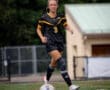 Female soccer player in action on the field, wearing a black jersey and knee brace, with a soccer ball in motion.
