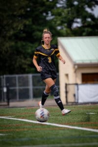 Female soccer player in action on the field, wearing a black jersey and knee brace, with a soccer ball in motion.