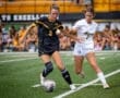 Two female soccer players compete for the ball on a field during a match, with spectators in the background.