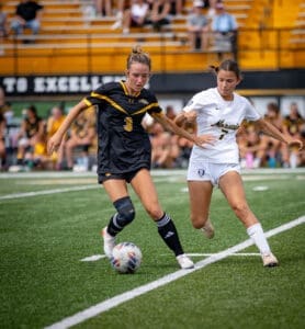 Two female soccer players compete for the ball on a field during a match, with spectators in the background.