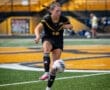 A young female soccer player in a black uniform dribbles a ball on a field with yellow bleachers behind her.