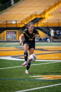 A young female soccer player in a black uniform dribbles a ball on a field with yellow bleachers behind her.