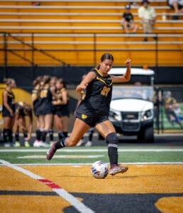 A female soccer player kicks a ball on the field during a sunny game, with teammates in the background.