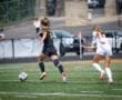 Two female soccer players chase a ball on the field during an intense match in bright summer sunshine.