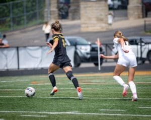 Two female soccer players chase a ball on the field during an intense match in bright summer sunshine.