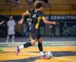 Young soccer player in a black uniform dribbles a ball on a brightly lit field during a night game.
