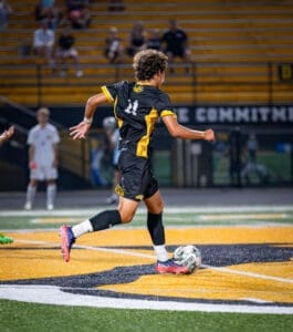 Young soccer player in a black uniform dribbles a ball on a brightly lit field during a night game.
