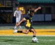 Soccer player in a black and yellow uniform strikes a ball on the field during a night game.