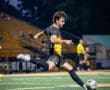 A soccer player in a black and yellow uniform strikes the ball on a well-lit field during a night game.