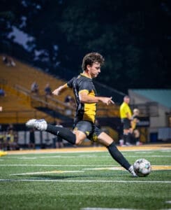 A soccer player in a black and yellow uniform strikes the ball on a well-lit field during a night game.