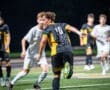 A soccer player in a black and yellow uniform dribbles the ball under night lights during an intense match.