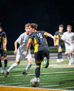 A soccer player in a black and yellow uniform dribbles the ball under night lights during an intense match.