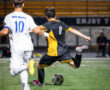 Player in black and yellow kicks a soccer ball on a field, with an opposing player in white nearby. Night game action shot.