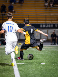 Player in black and yellow kicks a soccer ball on a field, with an opposing player in white nearby. Night game action shot.