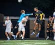 High school soccer players compete under the night sky, with a goalie leaping to block a ball aimed at the goal.