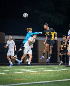 High school soccer players compete under the night sky, with a goalie leaping to block a ball aimed at the goal.