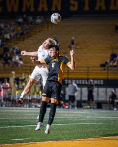 High school soccer players competing for the ball in mid-air at Newman Stadium during an evening match.
