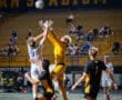 Goalkeeper leaps to catch the ball during an intense soccer match under stadium lights, with players competing for possession.