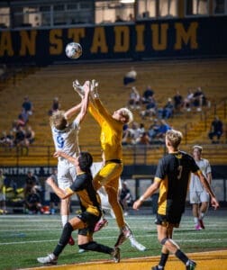 Goalkeeper leaps to catch the ball during an intense soccer match under stadium lights, with players competing for possession.