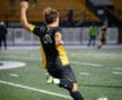 A soccer player in a black and yellow jersey takes a shot on goal during a night game on a turf field.