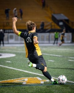 A soccer player in a black and yellow jersey takes a shot on goal during a night game on a turf field.