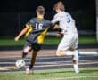 Two soccer players compete for the ball under nighttime lights, one wearing a black jersey with yellow stripes and the other in white.