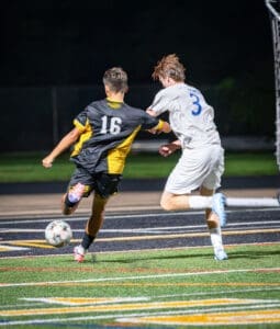 Two soccer players compete for the ball under nighttime lights, one wearing a black jersey with yellow stripes and the other in white.
