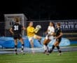 Goalkeeper catches soccer ball during a nighttime match, surrounded by players in action on the field.