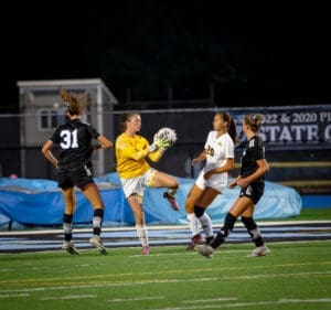 Goalkeeper catches soccer ball during a nighttime match, surrounded by players in action on the field.