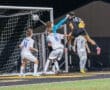 Goalkeeper leaps to block a shot during a high-stakes soccer match under the lights. Players compete for the ball.