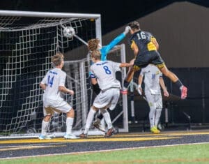 Goalkeeper leaps to block a shot during a high-stakes soccer match under the lights. Players compete for the ball.