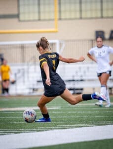 Soccer player in black jersey kicks a ball on a grass field during an intense match, with a teammate in the background.