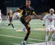 Player in a black soccer jersey leaps to control a ball during a night game on a turf field, with opposing players nearby.