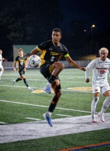 Player in a black soccer jersey leaps to control a ball during a night game on a turf field, with opposing players nearby.