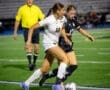 Two female soccer players compete for the ball during a night game, with a referee observing in the background.