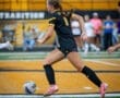 A soccer player in a black uniform races after the ball on a sunny field with cheering fans in the background.