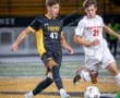 Two high school soccer players compete for the ball on the field during a night game, with a vibrant crowd in the background.