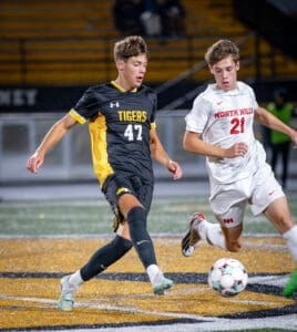 Two high school soccer players compete for the ball on the field during a night game, with a vibrant crowd in the background.