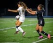 Two female soccer players compete for the ball under stadium lights during a night game on a turf field.