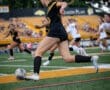 A female soccer player in black uniform races to kick the ball on a grassy field during a competitive match.