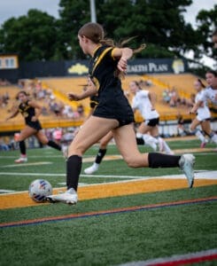 A female soccer player in black uniform races to kick the ball on a grassy field during a competitive match.