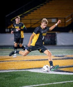 High school soccer player with jersey number 3 kicks a ball during a night game on a turf field.