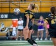 High school soccer player wearing jersey #10 jumps to head the ball, with fans cheering in the stands behind her.