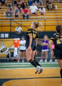 High school soccer player wearing jersey #10 jumps to head the ball, with fans cheering in the stands behind her.