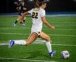 A soccer player in a white jersey #22 strides energetically to kick the ball on a well-lit field at night.