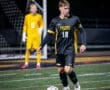 High school soccer player #18 dribbling a ball on the field with a goalkeeper in the background during a night game.