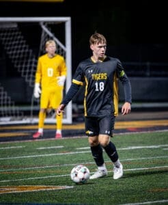 High school soccer player #18 dribbling a ball on the field with a goalkeeper in the background during a night game.