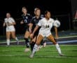 High school girls’ soccer match at night, player in white jersey prepares to receive the ball with teammates behind her.