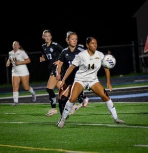 High school girls’ soccer match at night, player in white jersey prepares to receive the ball with teammates behind her.