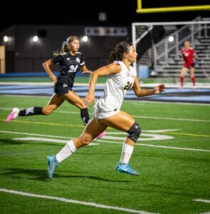 Two female soccer players sprinting on a field at night, showcasing athleticism and competition during a game.